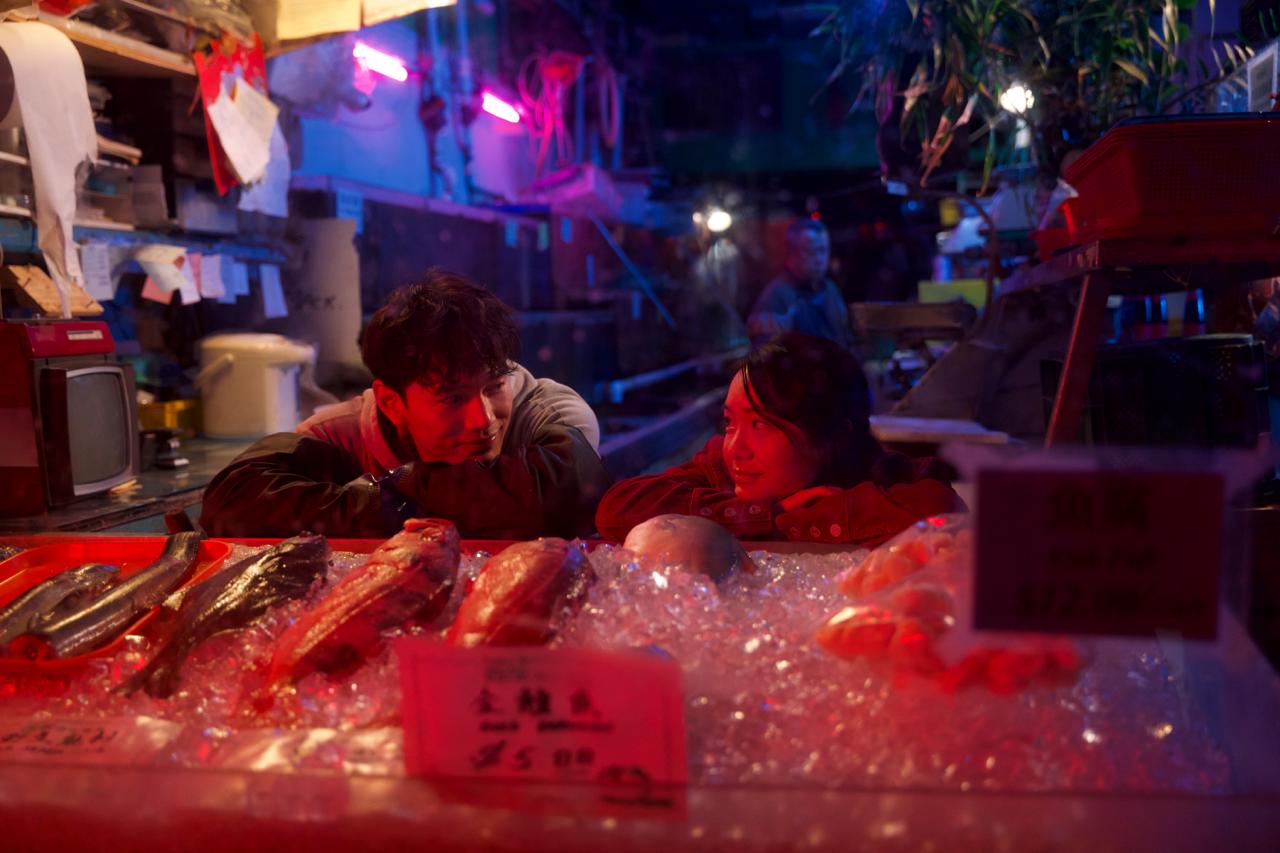 Two characters lean on a neon-lit fish-market counter